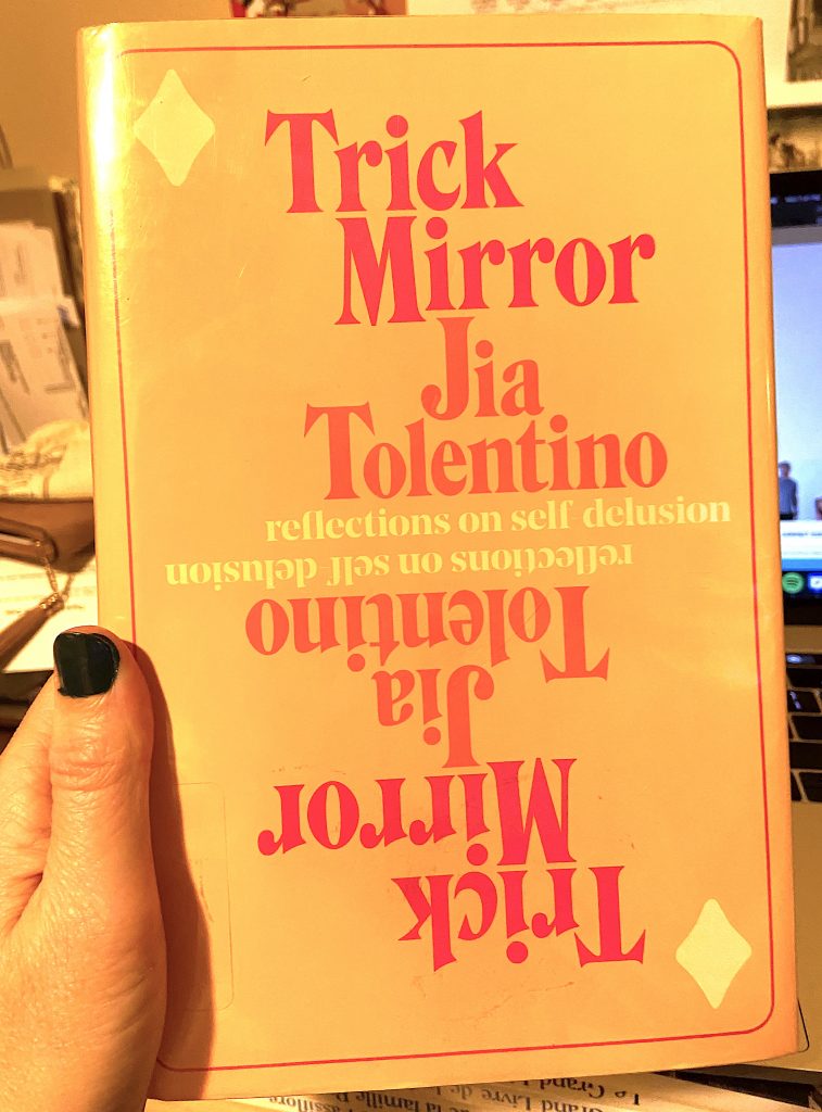 A hand holding a hardcover copy of Trick Mirror by Jia Tolentino. 